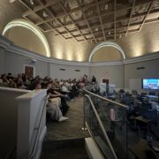 Photo of auditorium with audience and tv showing expert roundtable.