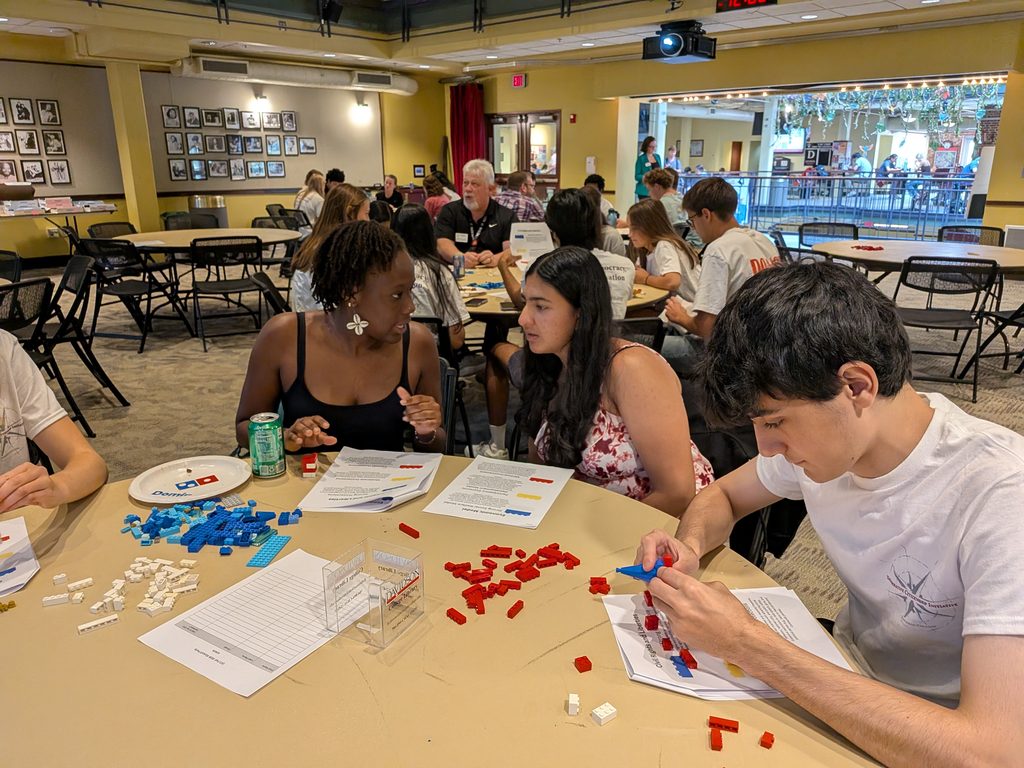 Students seated at a round table building with plastic bricks during a Deliberative Citizenship Initiative (DCI) interactive activity.