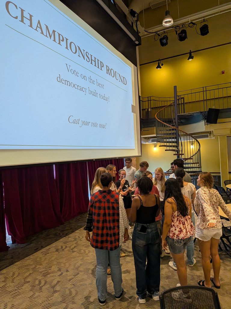 A group of students gathered around a large projection screen displaying voting instructions for the "Championship Round" during a Deliberative Citizenship Initiative (DCI) event.