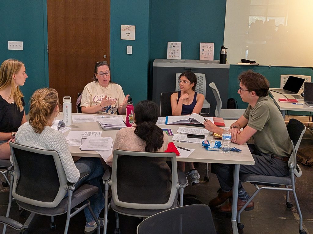 A group of five participants gathered around a table in a workshop room during a small-group deliberative dialogue session.